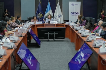 Un grupo de personas sentadas alrededor de una mesa de conferencias en forma de U, con banderas de Guatemala, pantallas, micrófonos y carteles de identificación