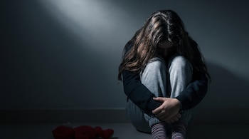 Adolescente sentada en el suelo con la cabeza baja, vestida con sudadera y medias de rayas. Junto a ella hay un oso de peluche rojo y una pared en penumbra.