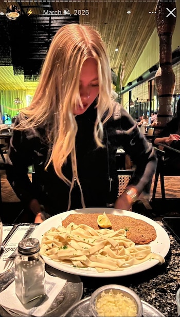 Mujer rubia mira un plato con milanesa, un gajo de limón y fideos. En la mesa hay un salero y un recipiente con queso rallado