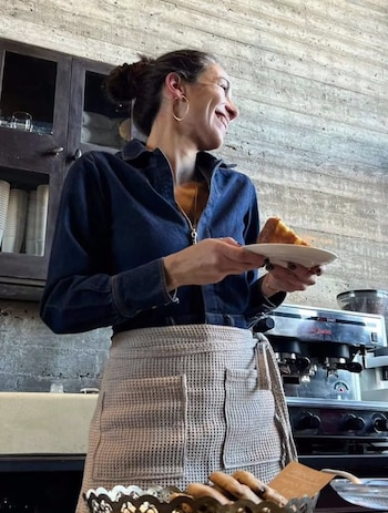 Una mujer con camisa de mezclilla y delantal sostiene un plato con una porción de torta junto a una cafetera