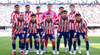 Once futbolistas del Club Deportivo Guadalajara posan para una fotografía grupal en un campo de fútbol. Visten uniformes rojos y blancos, excepto un portero con camiseta rosa.