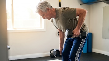 Un hombre con ropa deportiva flexiona el torso y levanta mancuernas en una sala con una ventana y accesorios de entrenamiento.