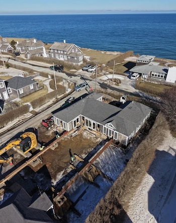 Vista aérea de una casa de madera gris siendo elevada con equipos de construcción, junto a una zanja de cimientos y trabajadores, cerca de la costa