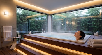 Mujer sumergida en un jacuzzi con vapor, sonriendo. Interior moderno con grandes ventanales a plantas verdes y madera. Toallas limpias y productos de baño a la vista.