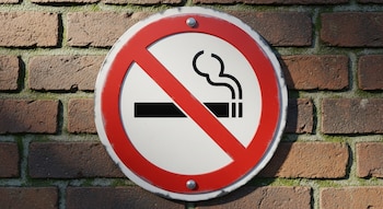 Un cartel circular blanco y rojo de 'prohibido fumar', fijado con tornillos, sobre una pared de ladrillo marrón rojizo con zonas de musgo verde.