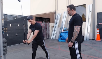 Un hombre vestido de negro realiza un ejercicio de tríceps en una máquina de cables al aire libre, mientras Jeff Cavaliere, con gafas y camiseta negra, lo observa