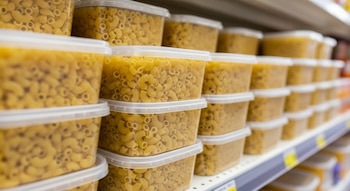 Múltiples envases plásticos transparentes llenos de macarrones con queso apilados en un estante de supermercado con un fondo desenfocado. No se observan marcas.