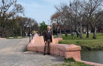 Un hombre mayor vestido con saco marrón y pantalón pardo está de pie sobre un puente rosado con barandillas negras, junto a un río y árboles