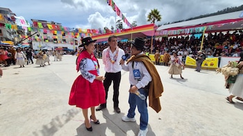 Un reportero con traje tradicional y micrófono entrevista a una mujer con vestido rojo y sombrero negro, junto a un hombre con camisa blanca, en un festival