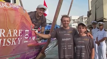 Tres hombres sonríen junto a un bote naranja y morado con 'Rare Whisky'. Uno está en el bote, dos de pie. Detrás, una bandera peruana y personal naval
