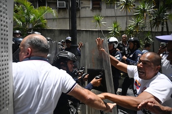 Agentes de seguridad con cascos y escudos transparentes se oponen a civiles. Palmeras y un edificio al fondo. Hombre con camiseta que tiene bandera de Venezuela