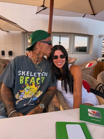 Hombre con gorra verde, gafas de sol y tatuajes besa la frente de una mujer con cabello oscuro, gafas de sol y blusa blanca en una terraza