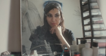 Una mujer con cabello oscuro, pañuelo y tatuajes se refleja en un espejo rodeado de frascos y organizadores de cosméticos