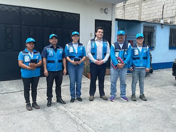 Seis personas con chalecos azules, gorras e identificaciones oficiales posan en fila frente a la entrada de una vivienda