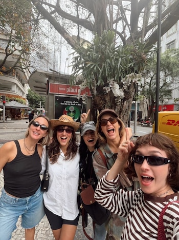 Cinco mujeres con gafas de sol sonríen y posan en una calle urbana frente a un árbol y un quiosco