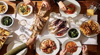 Vista superior de una mesa de madera con varias personas comiendo. Se ven platos de pasta, verduras asadas, pan, uvas, vino y un rallador de queso.