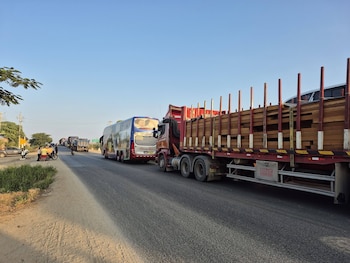 Una vista de una carretera con una larga fila de vehículos detenidos, incluyendo un autobús de pasajeros y varios camiones cargados de madera, bajo un cielo azul claro