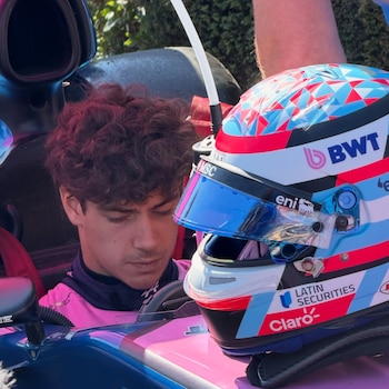 Hombre con cabello rizado castaño y traje rosa dentro de un monoplaza azul y rosa. A su lado hay un casco blanco, azul, rojo y rosa