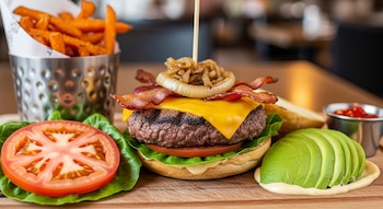Primer plano de una hamburguesa con queso, tocino, cebolla caramelizada y lechuga, acompañada de boniatos, aguacate, tomate y kétchup en una tabla.