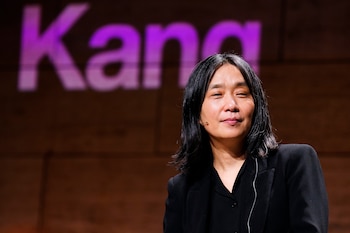Primer plano de una mujer asiática con cabello oscuro y blazer negro, sonriendo, con un micrófono en la mejilla y la palabra "Kang" borrosa detrás