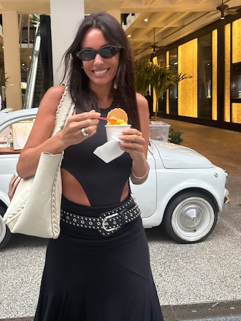 Pampita sonríe mientras come un helado en Miami, luciendo gafas de sol, un top negro, falda larga, cinturón de tachas y bolso blanco, con un coche Fiat 500 blanco detrás