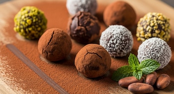 Nueve trufas de chocolate con diferentes coberturas de cacao, coco y frutos secos, junto a hojas de menta y granos de cacao sobre una tabla de madera.