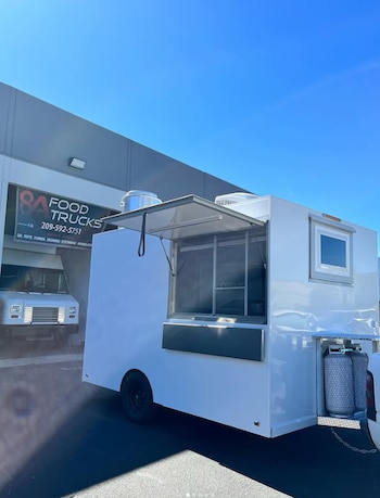 Un remolque blanco de comida rápida con la ventana de atención abierta está estacionado frente a un taller con el letrero 8A Food Trucks