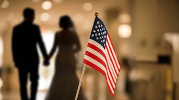 En primer plano, una bandera de Estados Unidos se muestra en detalle, mientras que en el fondo, dos siluetas borrosas, un hombre y una mujer, se toman de la mano.