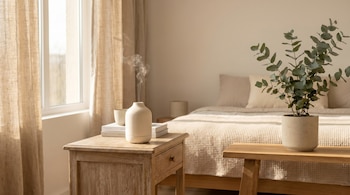 Dormitorio luminoso con cortinas claras, mesita de noche de madera, libros y un difusor de aromas. Una planta de eucalipto en maceta a la derecha.