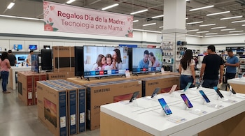 Clientes en una tienda de electrónica observan televisores y smartphones. Se ven pilas de cajas de televisores Samsung y un cartel de "Regalos Día de la Madre".