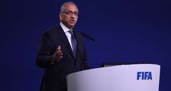 Hombre de traje y gafas hablando en un podio blanco con logo de FIFA azul, con un micrófono, sobre fondo azul oscuro