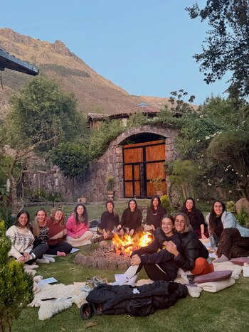 Un grupo de mujeres sentadas en el pasto alrededor de una fogata, con un edificio de piedra y puertas de madera y una montaña al fondo