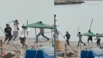 Varios hombres sostienen palos largos y se enfrentan en una playa de piedras cerca del mar y de embarcaciones pequeñas