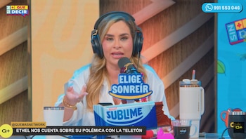 Una mujer con auriculares y micrófono frente a una mesa con una botella, una taza, una barra de chocolate y un cartel luminoso que dice Elige Sonreír