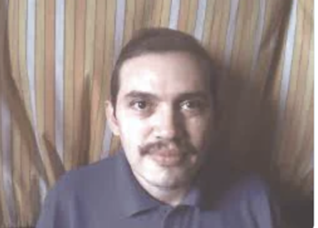 Hombre con bigote, camisa de cuello tipo polo, mirando al frente. Fondo con rayas verticales en tonos claros y oscuros