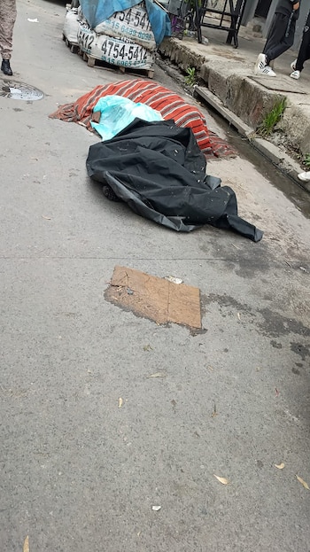 Un bulto cubierto con una lona negra, una bolsa azul y una manta tejida sobre el asfalto cerca del cordón de la acera