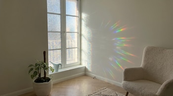 Rincón de una habitación con ventana de vidrio texturizado, reflejos con colores del arcoíris en la pared, un sillón y una planta en maceta.