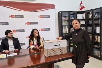 Una mujer deposita su voto en una urna de ONPE. Dos personas sonríen sentadas. Al fondo, logos de la Embajada de Perú en Qatar y estantes con libros. Bandera peruana visible