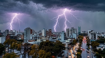 Panorámica urbana de edificios y calles mojadas bajo un cielo oscuro iluminado por descargas eléctricas.