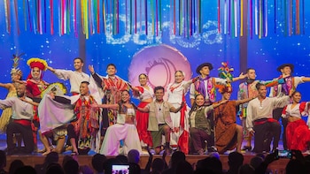 Diecinueve bailarines con diversos vestuarios tradicionales de Perú posan en un escenario con fondo azul, cintas de colores y una imagen circular; público en primer plano