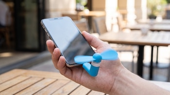 Una mano sujeta un teléfono móvil. Un miniventilador azul con aspas giratorias está conectado al puerto. Mesas y sillas de madera en el fondo borroso.