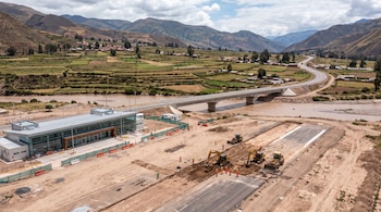 Una obra de construcción con maquinaria pesada, obreros, un edificio de vidrio, un puente sobre un río y montañas al fondo