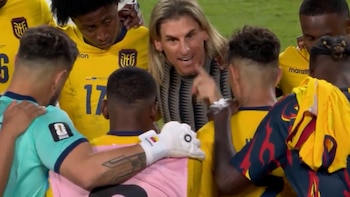 Hombre con cabello largo rubio habla a grupo de jugadores de fútbol con camisetas amarillas y una celeste, uno de ellos usa guante de portero blanco