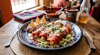 Plato de talavera con tres enchiladas rojas, queso, cebolla, frijoles refritos con totopos, lechuga, rábanos, cubiertos y una botella de refresco.