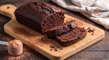 Un panqué de chocolate oscuro con dos rebanadas sobre una tabla de madera, acompañado de cacao en polvo, nibs y una cuchara con polvo.