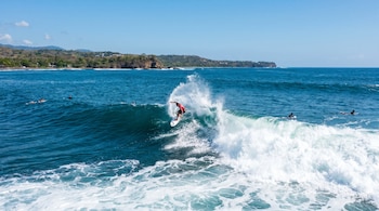 Surfista con camiseta roja en una ola rompiente con mucha espuma en el océano azul. La costa con vegetación y cielo despejado se ve al fondo, con otros surfistas cerca.