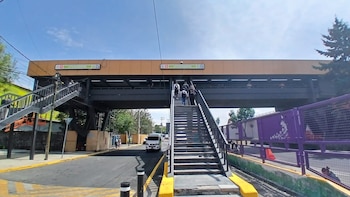 Vista exterior del Metro Universidad con escaleras oscuras llevando a una pasarela elevada. Personas suben y bajan. Hay una calle abajo y un cielo azul