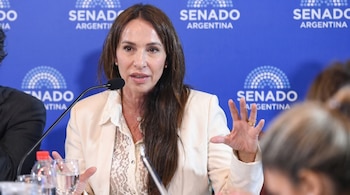 Mujer con cabello castaño largo, blazer crema y blusa de encaje blanco, hablando en un micrófono con el logo "Senado Argentina" en un fondo azul.