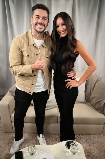 Nicolás Magaldi y Pampita de pie en un estudio, él con chaqueta beige y ella con vestido negro, ambos sonriendo frente a un sofá y cortinas blancas
