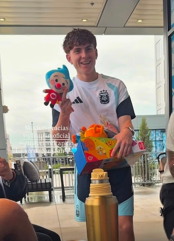 Un hombre joven sonríe vistiendo una camiseta de fútbol de la selección argentina, sostiene un muñeco azul y rojo y una caja de juguetes multicolor